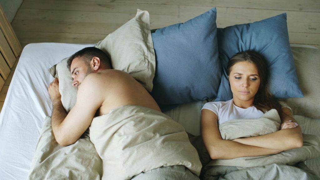 È bene parlare di sessualità col proprio neurologo Un uomo e una donna sdraiati a letto, lui girato su un fianco le dà le spalle, lei con le braccia conserte